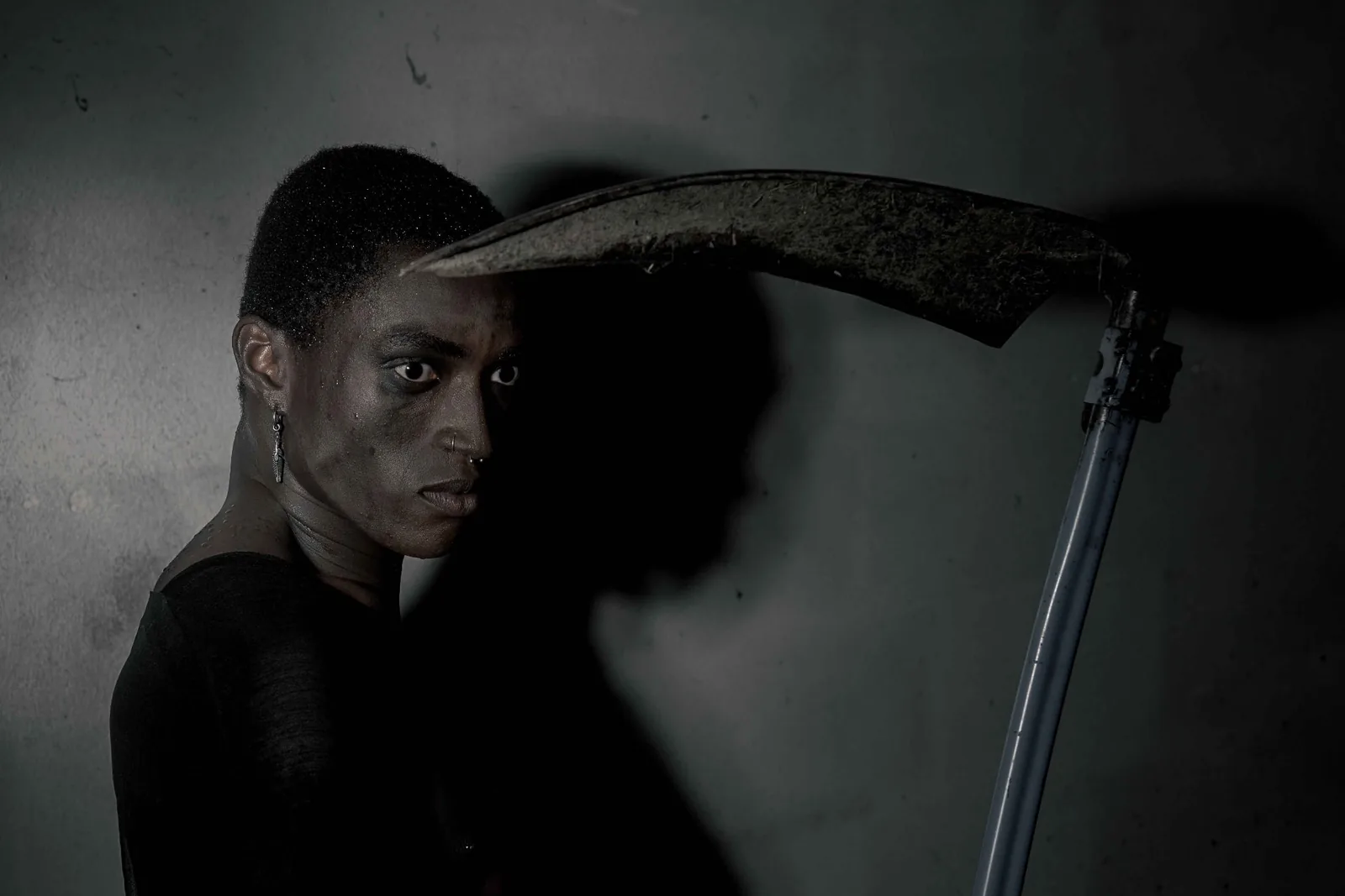 Dark editorial portrait of Kevin with a scythe, his shadow cast large on a concrete wall.