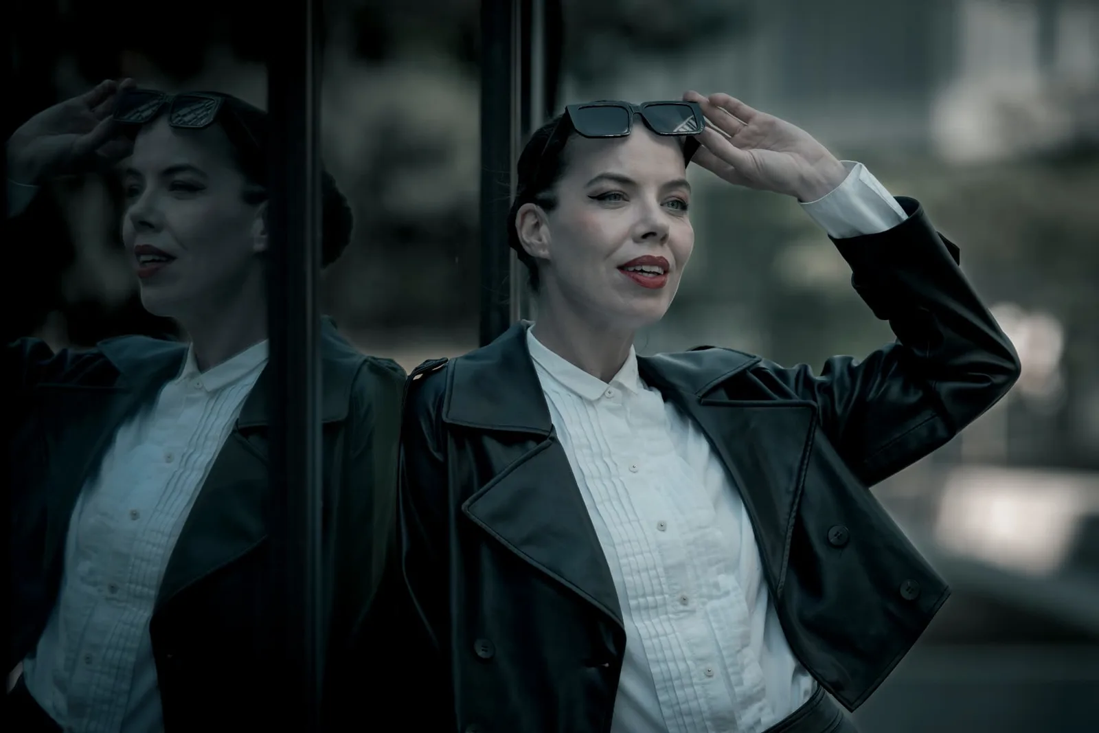 Denisa Strakova in a black leather coat and white shirt against a dark glass facade in Vienna, her full reflection standing beside her.