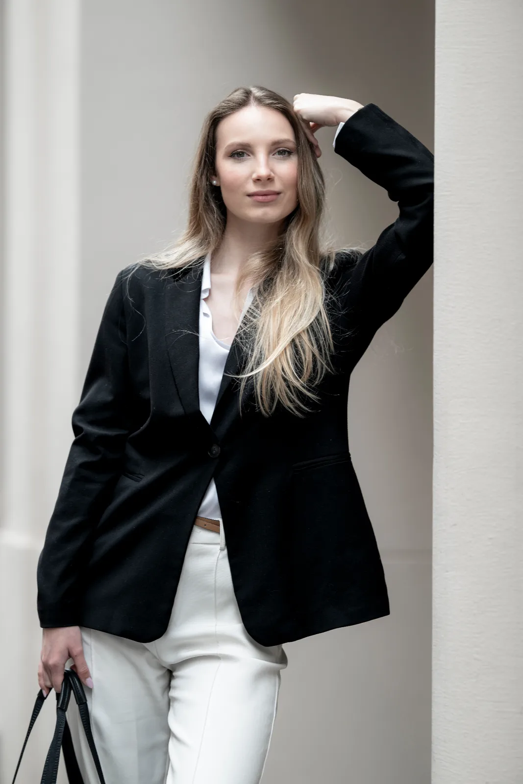 Full-length editorial portrait of a female executive in a black blazer and white trousers leaning against a column — confident leadership presence, business portrait photography Vienna Austria