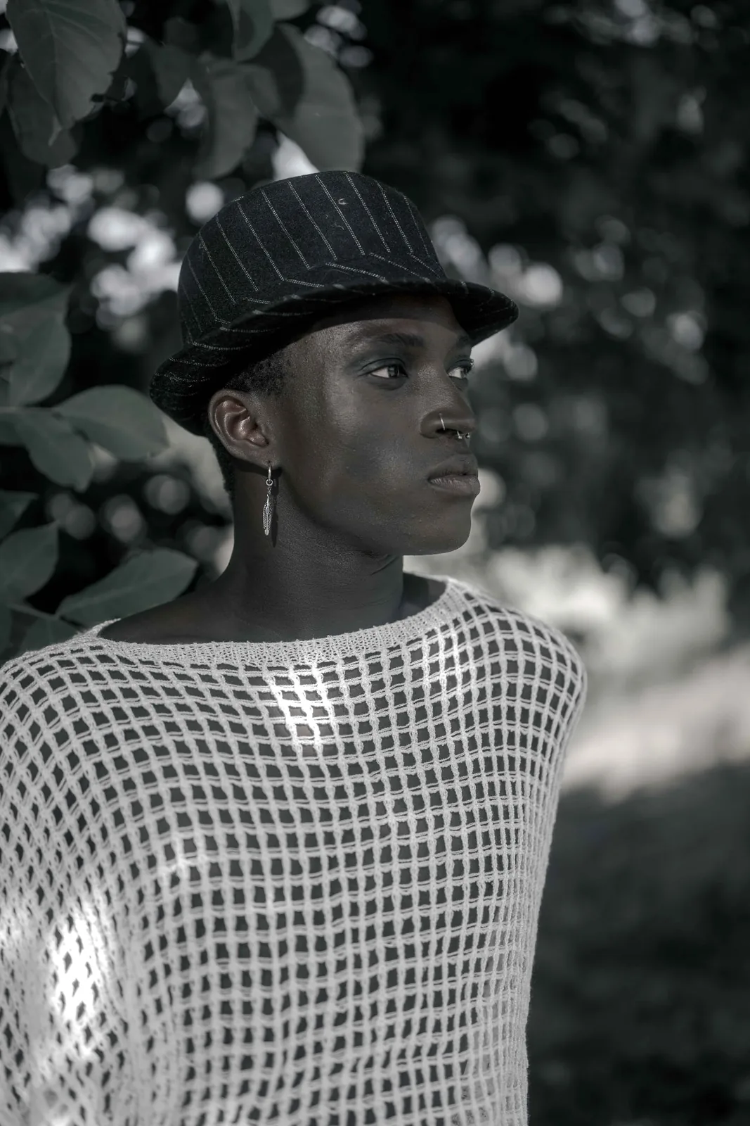 Male fashion editorial: Kevin wearing a mesh top and hat, photographed in reduced studio light.