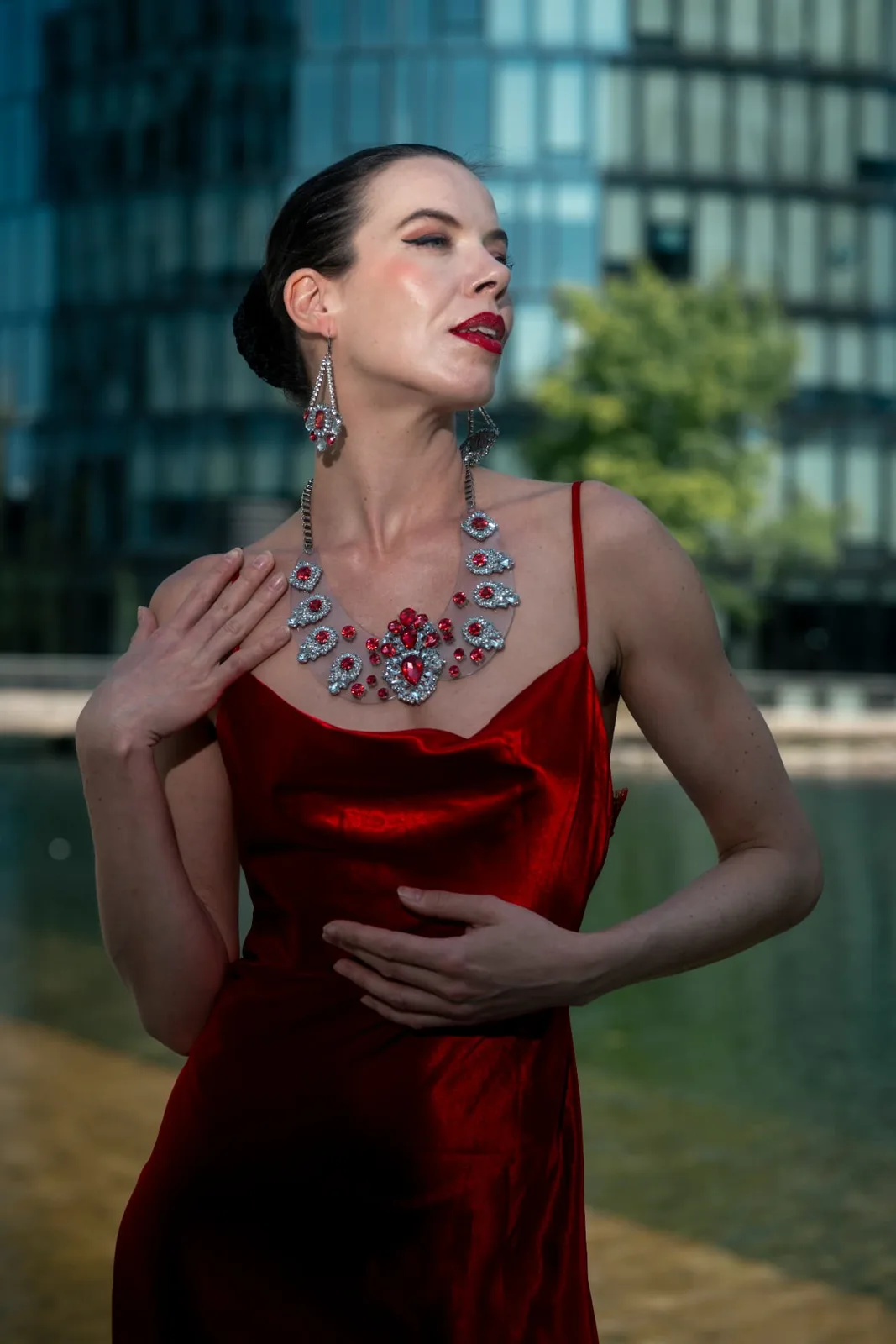Model Denisa Strakova in a red satin gown with a crystal statement necklace, separated from the glass facade behind her by a Godox AD200 Pro flash.