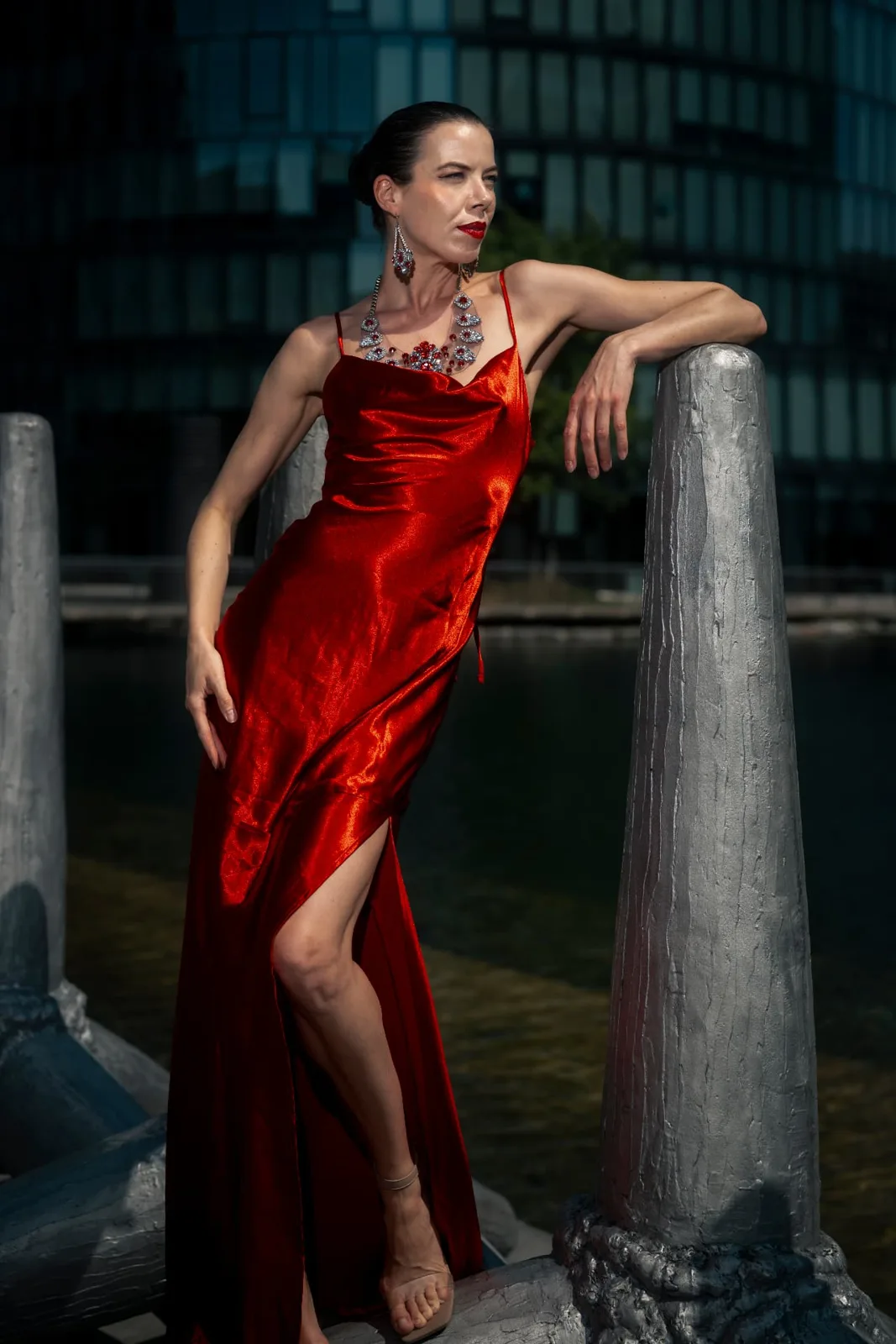 Model Denisa Strakova leaning against a concrete pillar at the water basin near Messe Wien, red gown sculpted by hard directional flash.