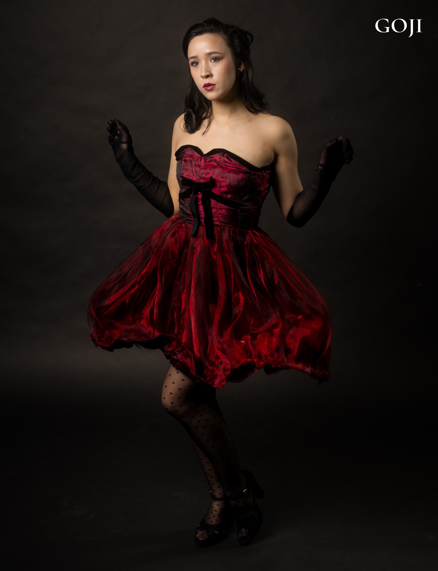 Crimson Poise editorial — GOJI Magazine Issue No. 187 — full-length fashion portrait of Victoria in a crimson velvet and organza dress with sheer black gloves, dark studio backdrop, photographed in Vienna by Peter Scholz