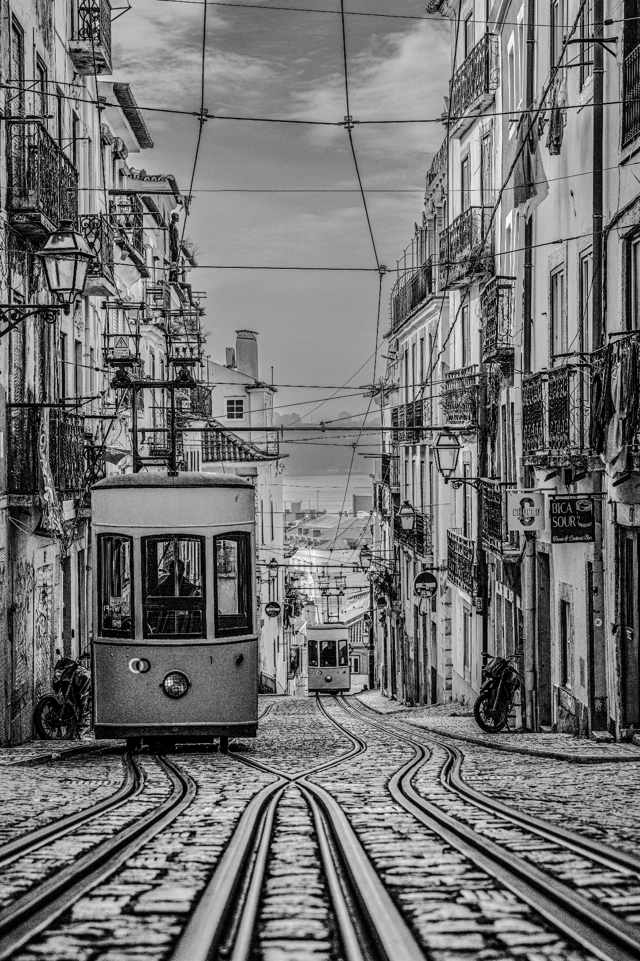 Two Elevador da Bica funicular cars meeting on steep street with Tagus river view — black and white fine art by schopho